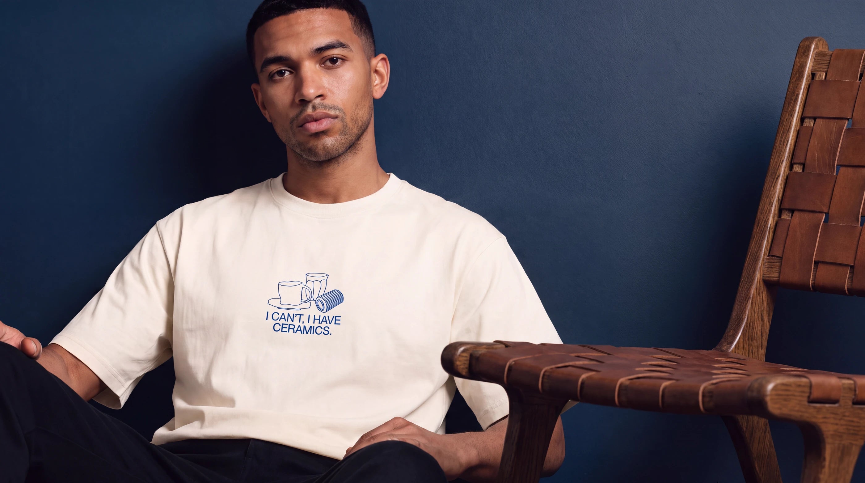 Model wearing the I Can't, I Have Ceramics T-shirt next to a woven leather chair. Image created or edited with AI.