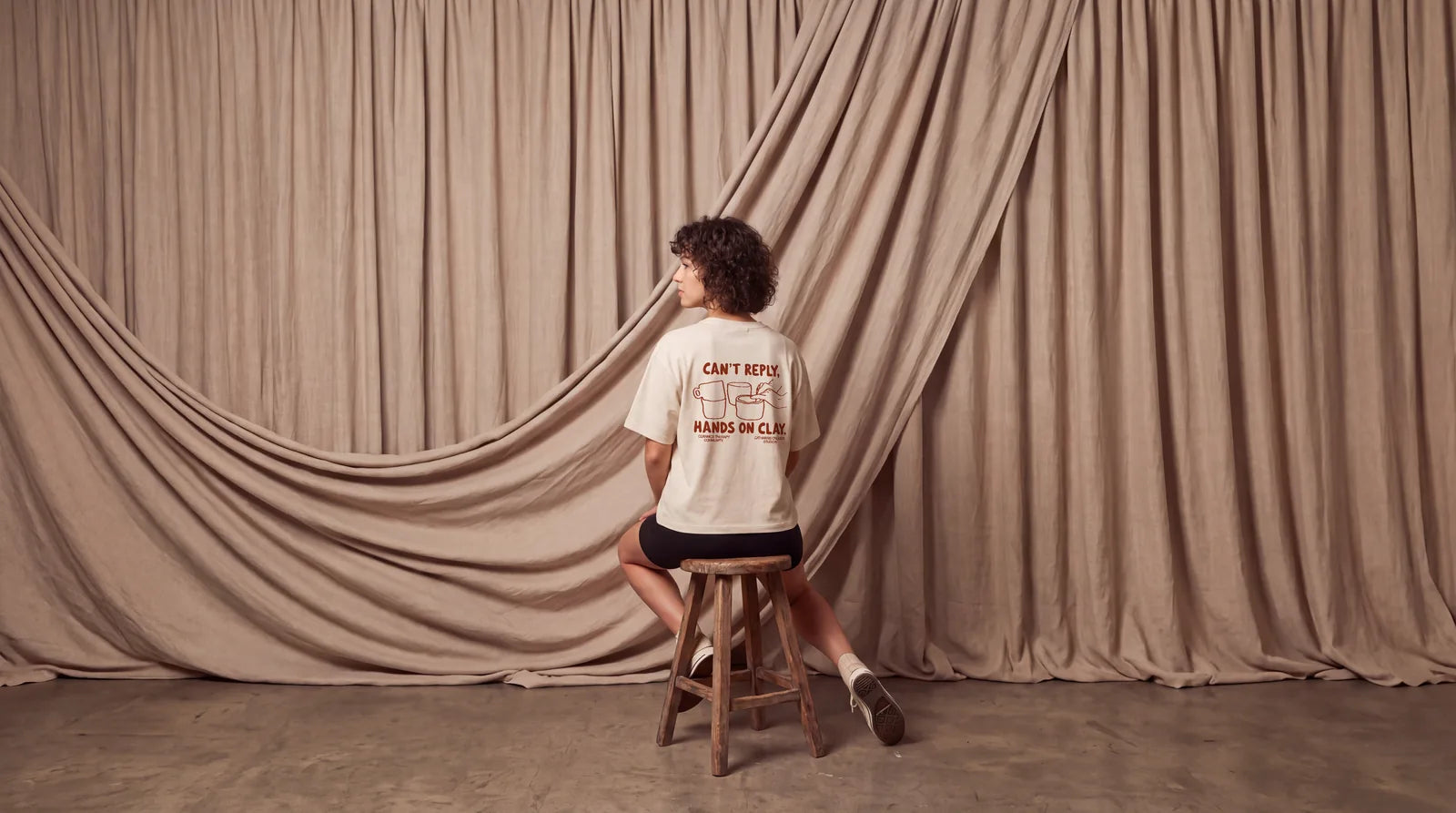 A woman with short curly hair sits on a wooden stool in front of a linen curtain, back to the camera, wearing the Can't Reply cropped T-shirt. Image created or edited with AI.