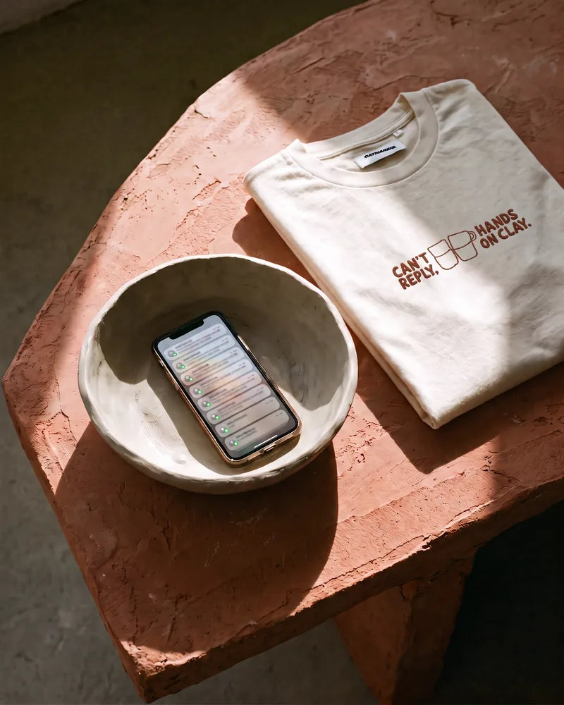The Can't Reply cropped T-shirt folded on a terracotta plaster table, next to a phone left inside a raw clay bowl, in hard sunlight. Image created or edited with AI.