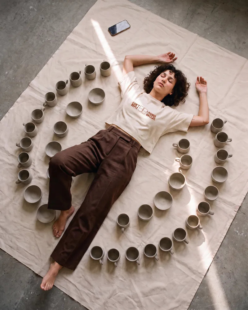 A woman with short curly hair lies on a raw canvas, eyes closed, inside a spiral of unfired mugs, her phone far out of reach. Image created or edited with AI.