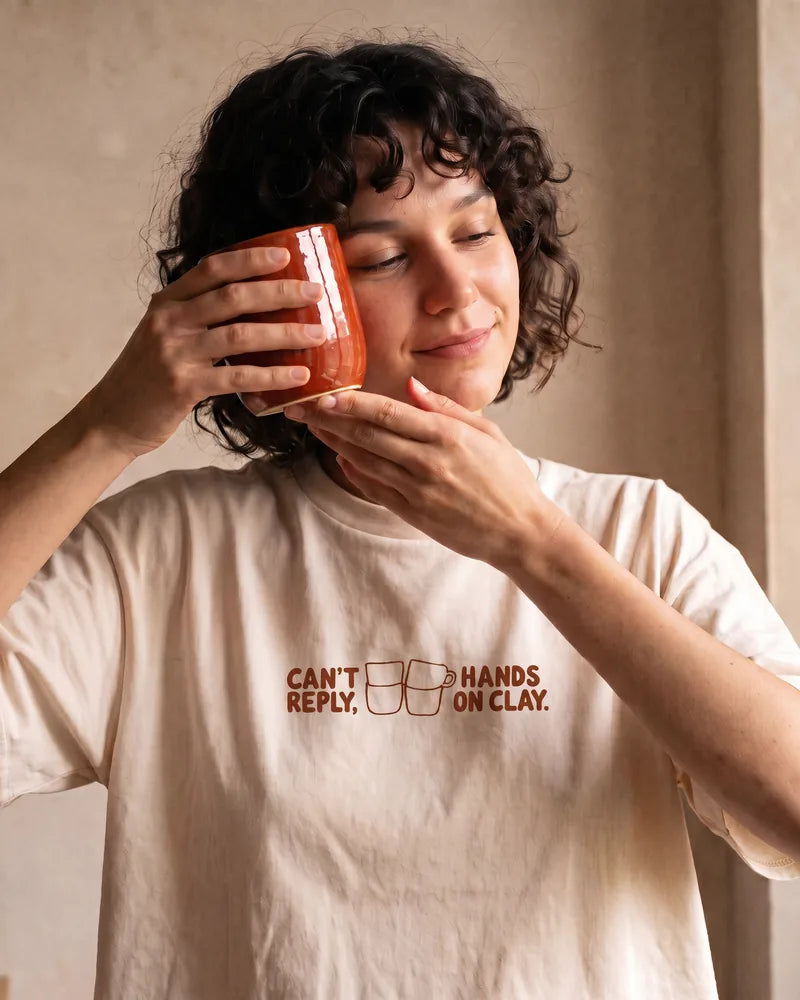 The woman holds a freshly glazed mug against her cheek and smiles, wearing the Can't Reply cropped T-shirt. Image created or edited with AI.