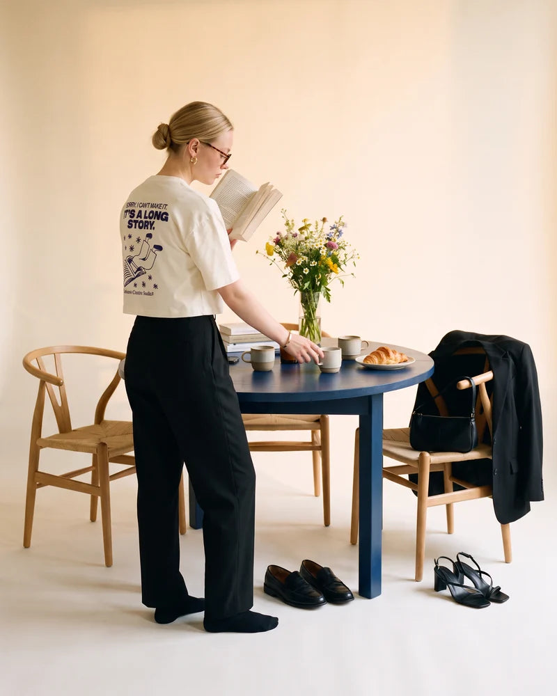 Woman standing by the table, reading while reaching for a mug without looking, wearing the cropped T-shirt Sorry, I can't make it. It's a long story. AI-generated image.