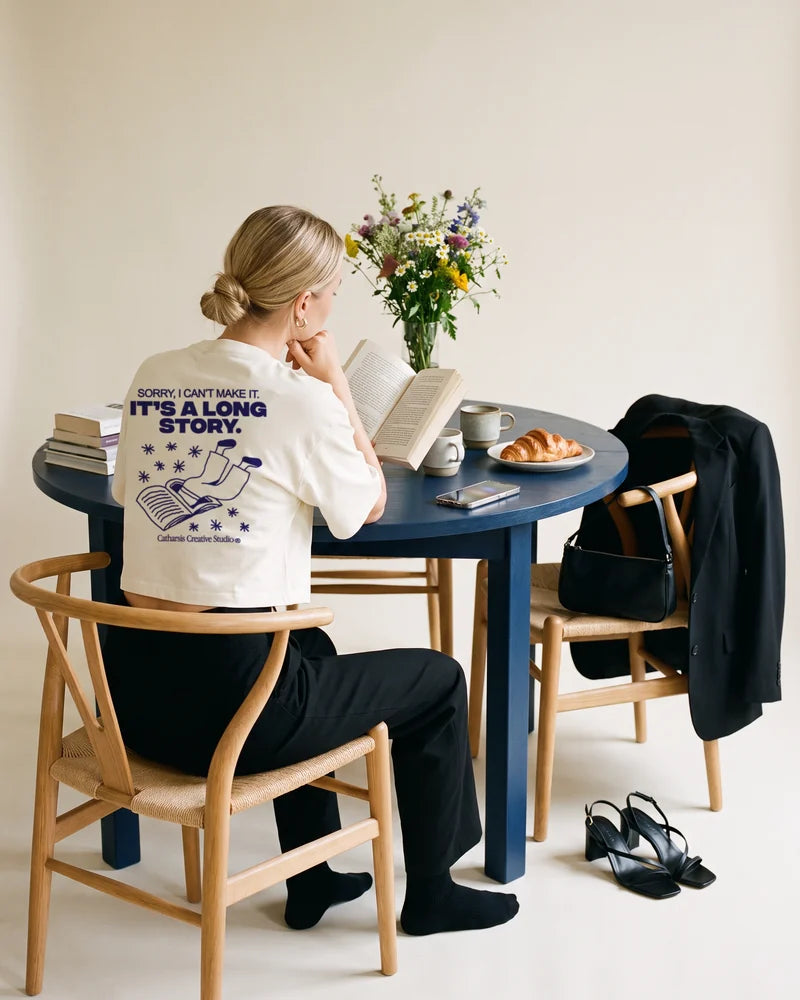Woman seen from behind, reading at midday next to an untouched croissant, wearing the cropped T-shirt Sorry, I can't make it. It's a long story. AI-generated image.