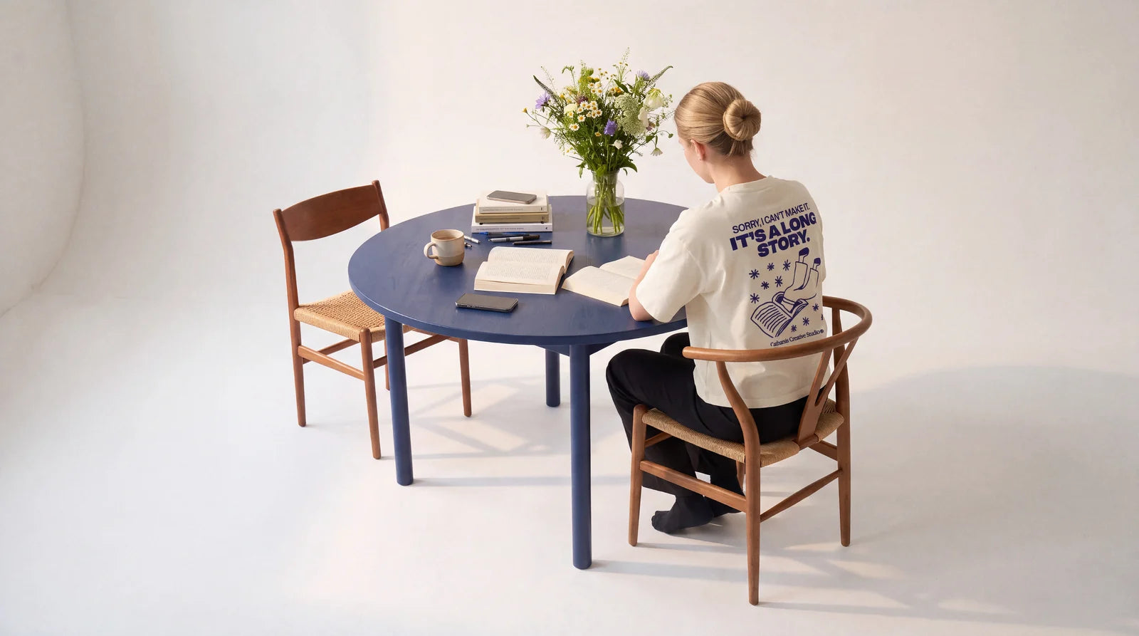 A woman reads at a round blue table, seen from behind, wearing the Sorry, I can't make it. It's a long story. T-shirt. Image created or edited with AI.