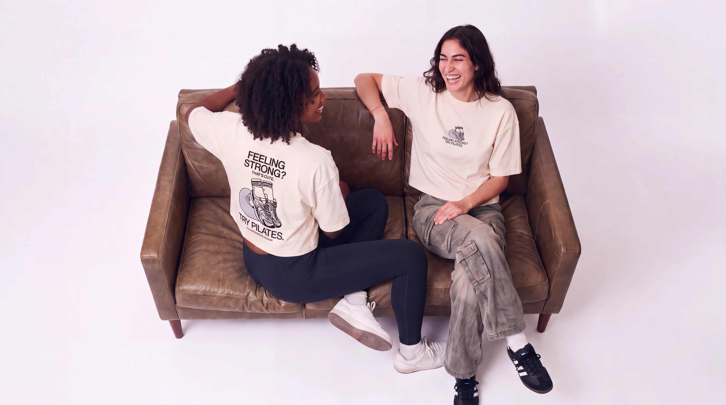 Two models laughing on a leather sofa in the Feeling Strong? That's Cute. Try Pilates cropped T-shirt. Image created or edited with AI.